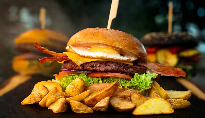 A hamburger with crispy bacon, fresh lettuce, and ripe tomato, served on a stylish black slate plate with grilled potatoes on the side.