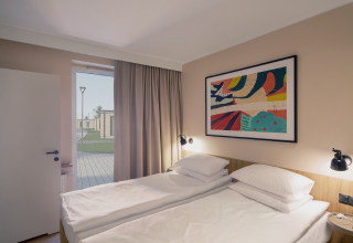 Hotel room with two single beds, modern lighting, and a colorful painting on the wall; an open door reveals a patio