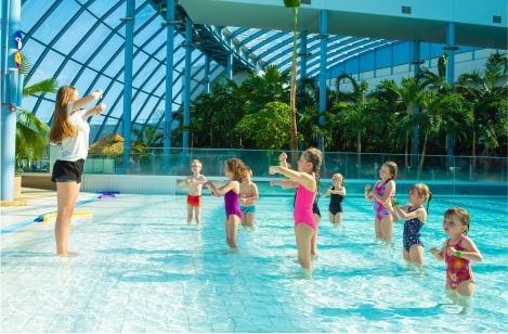 An instructor leads movement exercises in a shallow pool with a group of children, surrounded by tropical indoor waterpark scenery.