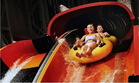 Two people sliding down a yellow raft waterslide in an indoor waterpark, with vivid slide colors in the background.