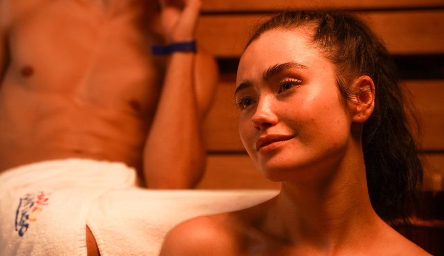 A man and woman sit side by side in a sauna, surrounded by wooden benches and steam, enjoying a moment of relaxation.